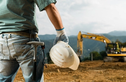 Im Vordergrund ist ein Bauarbeiter mit einem Helm in seiner Hand zu erkennen. im Hintergund sieht man einen Bagger.&nbsp;&copy;&nbsp;Fotolia.com