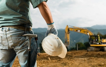Im Vordergrund ist ein Bauarbeiter mit einem Helm in seiner Hand zu erkennen. im Hintergund sieht man einen Bagger.