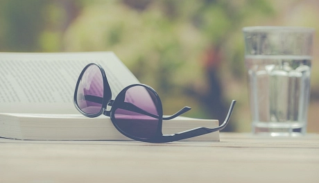 Das Bild zeigt ein aufgeschlagenes Buch im Sonnenlicht, eine Sonnenbrille und ein Glas Wasser.